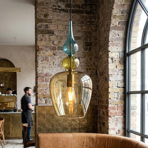 Puede incluir: Interior de un café moderno con una lámpara colgante de cristal ámbar en forma de lágrima. El espacio incluye una mesa de madera, asientos de cuero y una pared de ladrillo. Hay una taza de café y una planta suculenta sobre la mesa. La mesa mide unos 150 cm de largo.