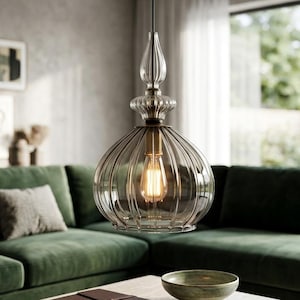 May include: A clear glass pendant light with a ribbed, globe-shaped shade and a decorative top. The light fixture hangs from a gold-coloured canopy and cord. An Edison-style bulb is visible inside the shade. The light is suspended above a coffee table in a living room.