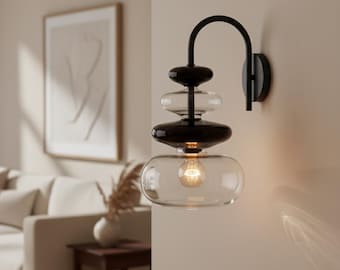 Könnte beinhalten: Eine moderne Wandleuchte mit einem schwarzen Metallarm und einem geschichteten Glasschirm. Der Schirm hat klare und dunkle Glaselemente, mit einer Glühbirne unten. Die Leuchte ist an einer neutralfarbenen Wand montiert.