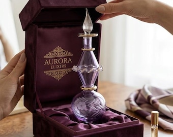 May include: A purple glass perfume bottle with a decorative stopper and gold accents, presented in a velvet box. The box is a deep purple color and has the words "AURORA ELIXIRS" in gold lettering. A small perfume vial is also visible.