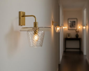 May include: A wall-mounted light fixture with a gold-colored metal arm and a clear, textured glass shade. The shade has a spiral design and is topped with a petal-shaped amber glass. The light is on, illuminating the hallway.