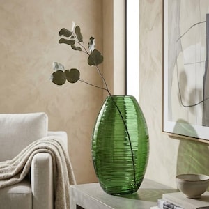 May include: A green glass vase with a ribbed texture, holding a sprig of greenery. The vase is oval-shaped and sits on a concrete table. Books and a small bowl are also on the table. An abstract artwork in a frame hangs on the wall.
