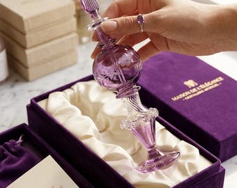 May include: A decorative, purple glass perfume bottle with a bulbous body and a long, ornate stem. The bottle is held above a purple velvet box with a white satin lining. The box contains a certificate of authenticity. The text "MAISON DE L'ÉLÉGANCE" is visible on the box.