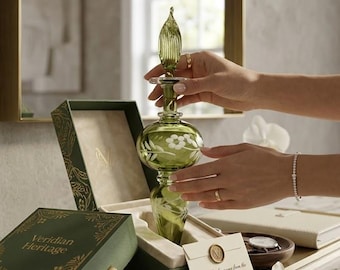 May include: A green glass perfume bottle with a floral design is held near its open box. The box and bottle are part of the Veridian Heritage Collection. The bottle has a decorative stopper and is displayed on a marble surface.