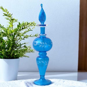 Op de afbeelding: Een blauwe glazen fles met een bloemenpatroon. De fles heeft een lange, slanke hals en een rond lichaam. De fles is versierd met een wit bloemenpatroon.