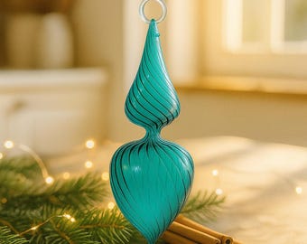 Puede incluir: Adorno navideño de cristal color turquesa con diseño en espiral y lazo transparente para colgar. El adorno está sobre una mesa de madera con palitos de canela y agujas de pino. El adorno mide aproximadamente 15 cm de alto.