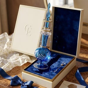 May include: A blue glass perfume bottle with a decorative stopper and gold accents, presented in a cream-colored box with a blue velvet interior. The box is tied with a blue ribbon. The bottle is approximately 6 inches tall.