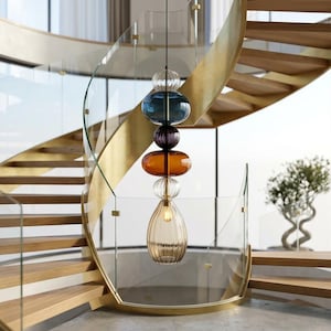 May include: A modern spiral staircase with wooden steps and a brass-colored handrail. A multi-colored glass pendant light hangs in the center. The staircase is surrounded by glass panels, and a small tree is visible.