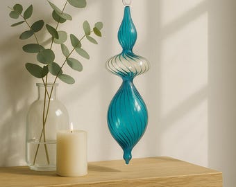 Puede incluir: Adorno de cristal turquesa con diseño en espiral colgado en la pared. Un jarrón de cristal transparente con hojas verdes y una vela encendida sobre una estantería de madera. El adorno tiene un lazo plateado en la parte superior.
