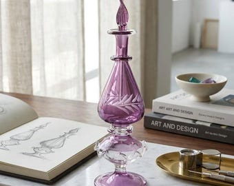 May include: A decorative, purple glass perfume bottle with a detailed stopper and etched leaf design. The bottle sits on a marble surface with a gold pen, a sketch book, and a small amethyst crystal. The text "PROTOTYPE PHASE VIGNETTE No. 1" is visible.
