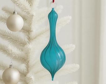 Puede incluir: Un adorno navideño de cristal color turquesa con líneas verticales y forma de lágrima cuelga de una cinta roja. También se ven dos bolas blancas y plateadas, colgadas de un árbol de Navidad blanco. Decoración navideña.