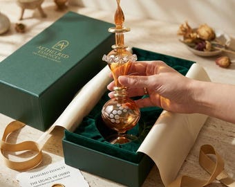 May include: An amber-colored glass perfume bottle with a decorative stopper is displayed in a green box. The bottle has a round base with a pattern of white dots. The box contains a card that reads "Aethelred Essences The Legacy of Osiris Handcrafted Artisanal Glass."