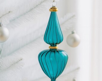 Puede incluir: Adorno navideño de cristal color turquesa con detalles dorados. El adorno tiene una forma única con una base bulbosa y una parte superior puntiaguda, con ranuras verticales. Cuelga de un hilo dorado sobre un árbol de Navidad blanco.