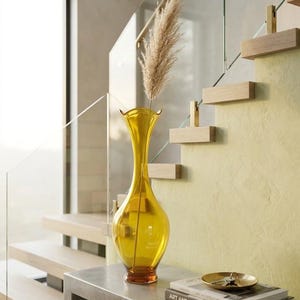 May include: A tall, yellow glass vase with a flared top and a brown base, holding a dried plant. The vase sits on a gray concrete table with a book titled "ART AND ARCHITECTURE" and a small gold dish. A staircase is in the background.