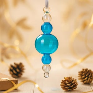 May include: A clear, turquoise glass ornament with a teardrop shape. The ornament has a series of rounded shapes, with a clear glass top and bottom. A silver ribbon is attached for hanging. The background includes pinecones and a gift box.