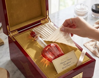 May include: A red glass perfume bottle with a decorative stopper inside a red wooden box with gold lining. The box is open, revealing the bottle and a cream-colored fabric. A label reads "Aethel - Masterpiece Collection - Seraphina".