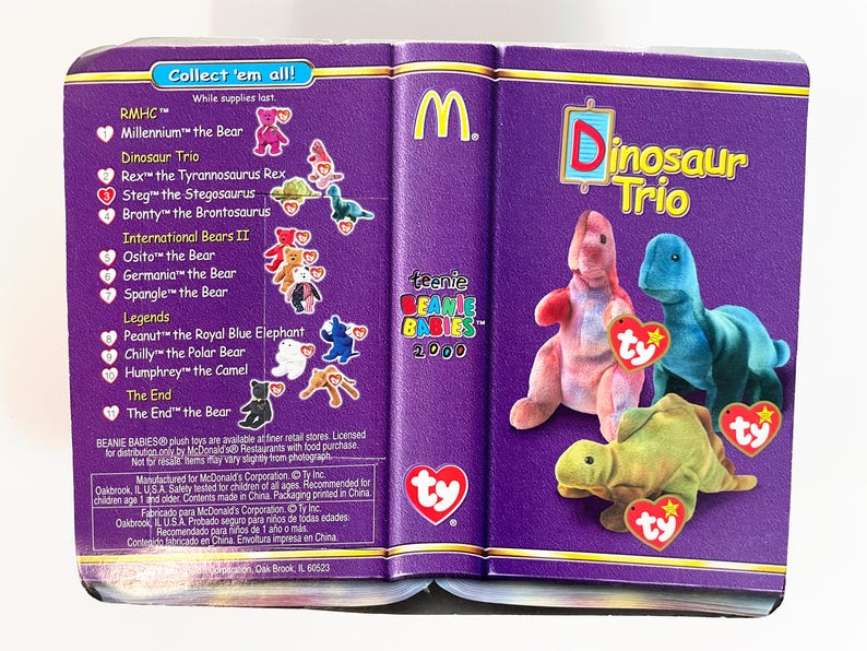 Pu&ograve; includere: Un contenitore viola a forma di libro con la scritta "Dinosaur Trio" e il logo McDonald's. Il contenitore contiene tre giocattoli di dinosauri di peluche con etichette a cuore Ty. Il lato sinistro elenca altri Beanie Babies.