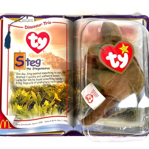 Pu&ograve; includere: Un peluche Ty Beanie Baby Steg il Stegosauro, ancora nella sua confezione originale. La confezione include un libretto con il testo "Dinosaur Trio" e la storia di Steg. Il peluche &egrave; marrone e ha un'etichetta a forma di cuore rossa.