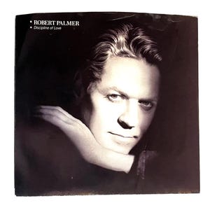 May include: Black and white album cover featuring Robert Palmer. The text "ROBERT PALMER" and "Discipline of Love" are printed at the top. Palmer's face is in the center, with his arm resting on his chin.