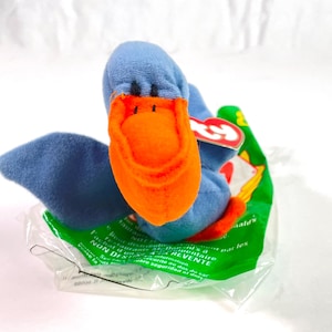May include: A blue plush duck toy with an orange beak and a red and white tag. The duck is sitting on a green and white McDonald's packaging.