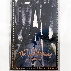 May include: A cassette tape with a black and white image of people standing. The text "The Wallflowers" is in gold script. The cassette case is clear with a gold border.