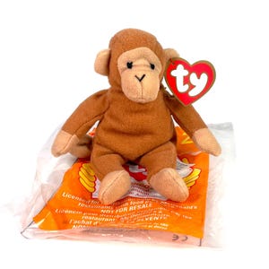 May include: A brown plush monkey toy with a red heart-shaped tag that says "ty" in white. The monkey is sitting on a clear plastic bag with orange and yellow graphics and text that reads "Licensed for restaurants with food promotions. NOT FOR RESALE."