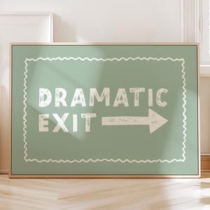 Op de afbeelding: Een ingelijst bord met de woorden "DRAMATIC EXIT" in witte blokletters en een pijl naar rechts op een saliegroene achtergrond. Het bord heeft een decoratieve witte rand en een houten frame. Het bord wordt binnenshuis weergegeven.