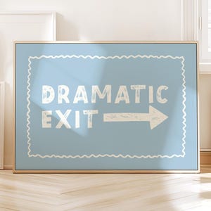 May include: A framed sign with a light blue background and white text that reads "DRAMATIC EXIT" with an arrow pointing right. The sign has a decorative white border and a wooden frame.
