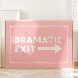 Op de afbeelding: Een roze ingelijst bord met de witte woorden "DRAMATIC EXIT" en een witte pijl naar rechts. Het bord heeft een decoratieve witte rand en wordt weergegeven in een lichte kamer.
