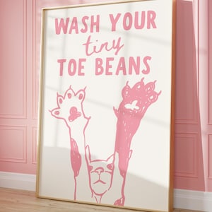 May include: A framed art print with the text "WASH YOUR tiny TOE BEANS" in pink. The artwork features a cat illustration with raised paws in pink. The background is white, and the frame is gold.