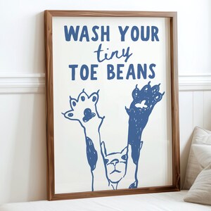 Puede incluir: Un estampado enmarcado con el texto "WASH YOUR tiny TOE BEANS" en azul. La obra de arte presenta huellas de patas azules y una ilustración de un perro. El marco es de madera y la impresión se muestra sobre un fondo blanco.