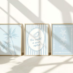 Op de afbeelding: Drie ingelijste kunstprints met een kustthema. De prints tonen een silhouet van een palmboom, de tekst "the world is your oyster" met een oesterafbeelding en drie zeesterren. De prints zijn in lichtblauw en wit.