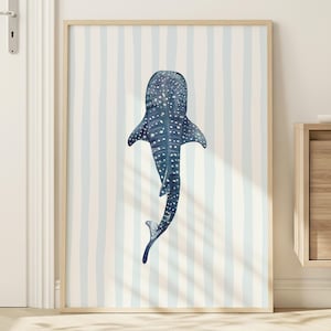 May include: A framed art print depicting a whale shark illustration in watercolour. The artwork uses blue and white tones, set against a striped background. The print is presented in a light wood frame, ideal for a coastal or nautical theme.
