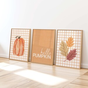 Puede incluir: Tres láminas decorativas en marcos de madera clara. La lámina izquierda muestra una calabaza naranja sobre un fondo de cuadros vichy beige. La central es de color naranja sólido con el texto blanco hello PUMPKIN. La derecha muestra tres hojas.