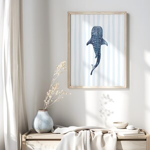 May include: A framed art print featuring a watercolor illustration of a whale shark. The artwork is in shades of blue and white, with a striped background. The print is displayed in a light-colored wooden frame, suitable for a coastal or nautical-themed decor.