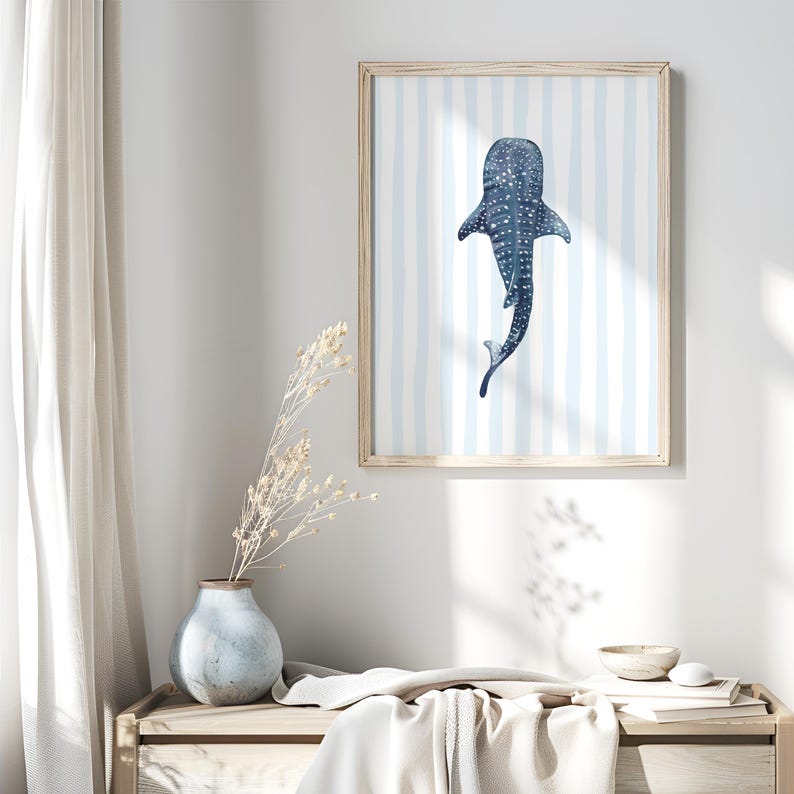May include: A framed art print featuring a watercolor illustration of a whale shark. The artwork is in shades of blue and white, with a striped background. The print is displayed in a light-colored wooden frame, suitable for a coastal or nautical-themed decor.