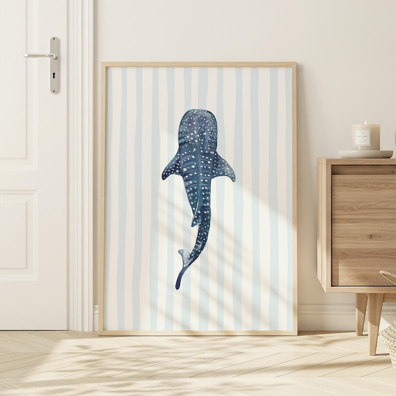 May include: A framed art print featuring a watercolor illustration of a whale shark. The artwork is in shades of blue and white, with a striped background. The print is displayed in a light wood frame, suitable for a coastal or nautical theme.