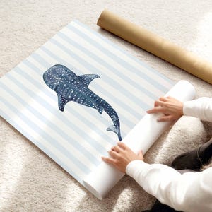 May include: A rolled-up print featuring a blue whale shark illustration with white spots, set against a backdrop of light blue and white stripes. The print is being unrolled on a light-colored rug.