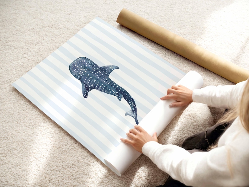 May include: A rolled-up print featuring a blue whale shark illustration with white spots, set against a backdrop of light blue and white stripes. The print is being unrolled on a light-colored rug.