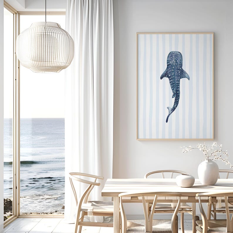May include: A framed art print featuring a blue whale shark illustration on a light blue and white striped background. The artwork is displayed in a bright, airy dining room setting with a view of the ocean.