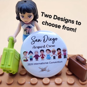 May include: A round pinback button featuring the text San Diego Airport Crew 2026 International Convention, an illustration of diverse people holding hands, and a small airplane. A plastic figure holds a green suitcase next to the button.