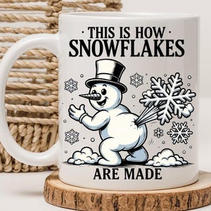 Puede incluir: Taza de cerámica blanca con un muñeco de nieve y el texto "THIS IS HOW SNOWFLAKES ARE MADE". El muñeco de nieve lleva un sombrero de copa y expulsa copos de nieve. La taza está sobre un posavasos de madera.