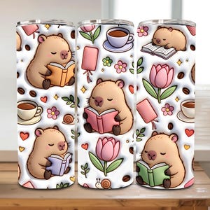 May include: Three insulated stainless steel tumblers with a repeating pattern of a cartoon capybara reading a book, coffee cups, flowers, and hearts. The background is white. Each tumbler is designed to keep drinks hot or cold.