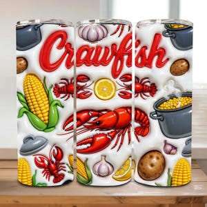 May include: A white tumbler with a vibrant design featuring the word "Crawfish" in red, along with illustrations of red crawfish, corn, potatoes, garlic, and lemon slices. The design includes cooking pots, suggesting a culinary theme.