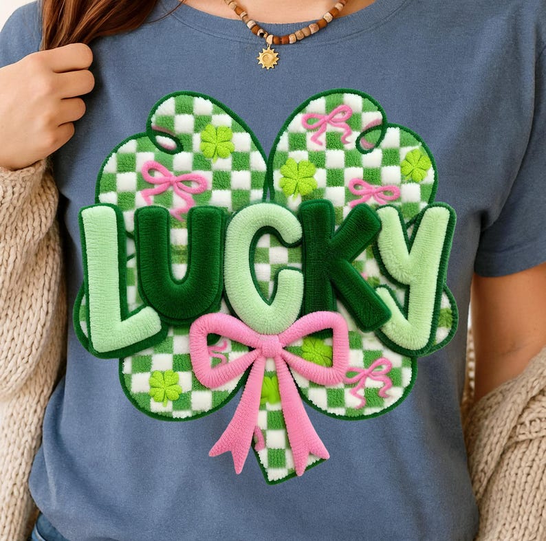 May include: A blue t-shirt featuring a green and white checkered shamrock design with pink bows and the word "LUCKY" in green and light green. The shirt is a St. Patrick's Day theme.