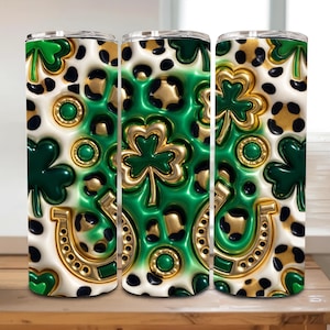 May include: Three stainless steel tumblers with a St. Patrick's Day theme. The design features green shamrocks, gold horseshoes, and leopard print spots on a white background. The tumblers are cylindrical and have a glossy finish.