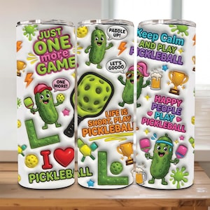 May include: Three white stainless steel tumblers with pickleball-themed graphics. The designs include cartoon pickles, pickleball paddles, and phrases like "Just One More Game" and "Keep Calm and Play Pickleball." The tumblers are decorated with bright colors.