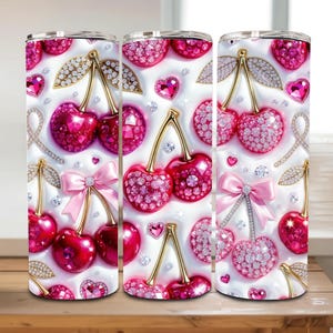 Puede incluir: Tres vasos de acero inoxidable con un patrón repetido de cerezas rosas, corazones y lazos sobre un fondo blanco. Las cerezas y los corazones están adornados con pedrería y detalles dorados. El diseño incluye detalles de diamantes.