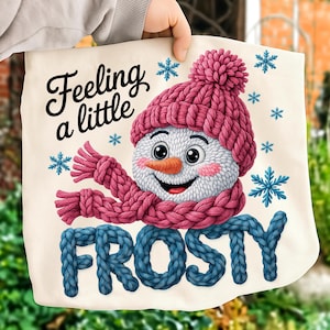 Puede incluir: Un bolso tote color crema con un muñeco de nieve sonriente que lleva un gorro y una bufanda de punto rosa. El texto "Feeling a little FROSTY" se muestra en letras de punto negras y azules, con copos de nieve azules.