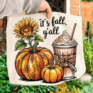 Puede incluir: Bolso tote color crema con una ilustración de girasol, calabazas y un café con leche de calabaza especiada. El texto "it's fall, y'all" está escrito en negro. Las calabazas son naranjas con diseños florales.